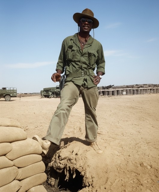 Leon Collier standing in the desert above a sandbagged bunker hole, wearing a bush hat and sunglasses.